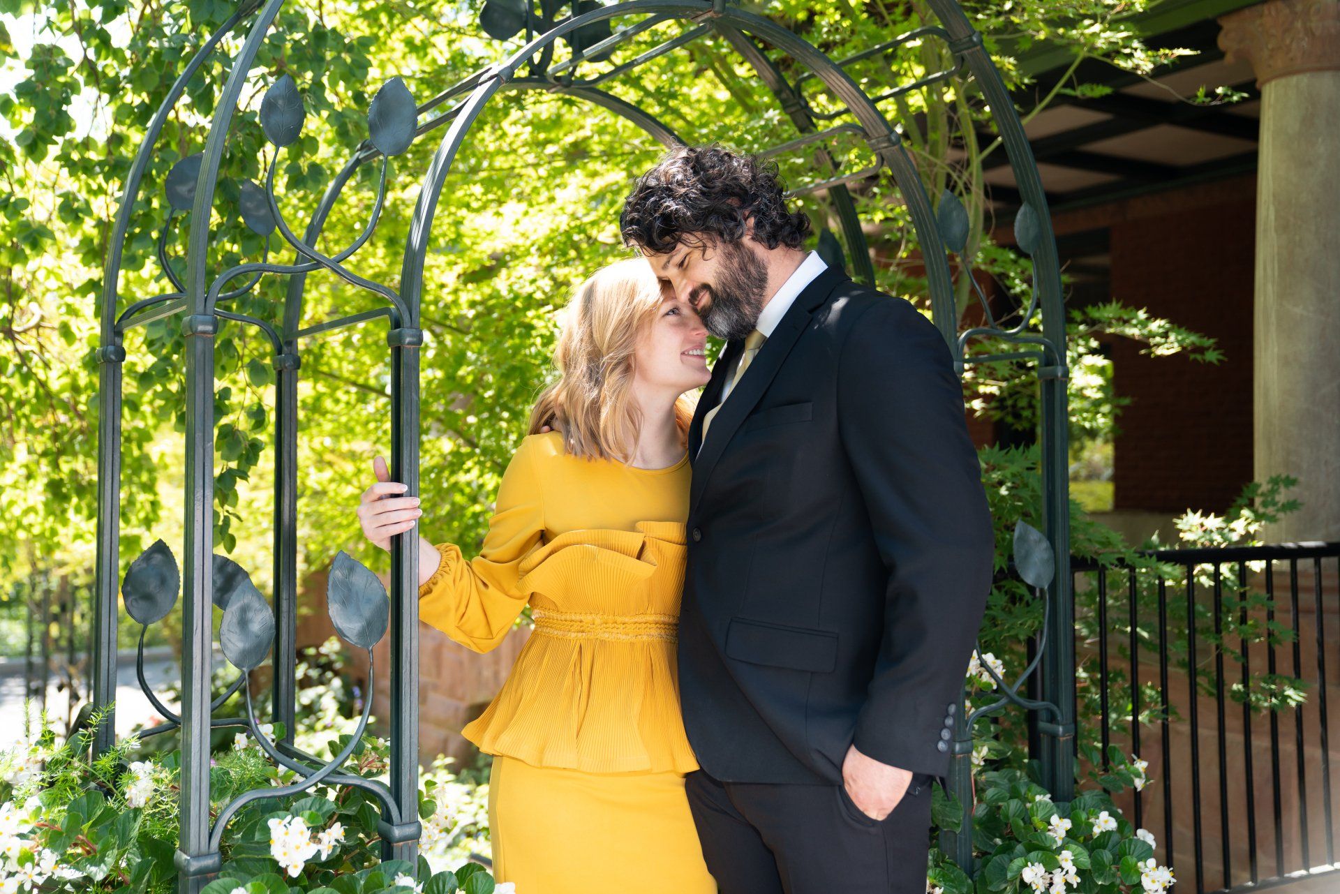 A man in a suit and a woman in a yellow dress are kissing under an archway.