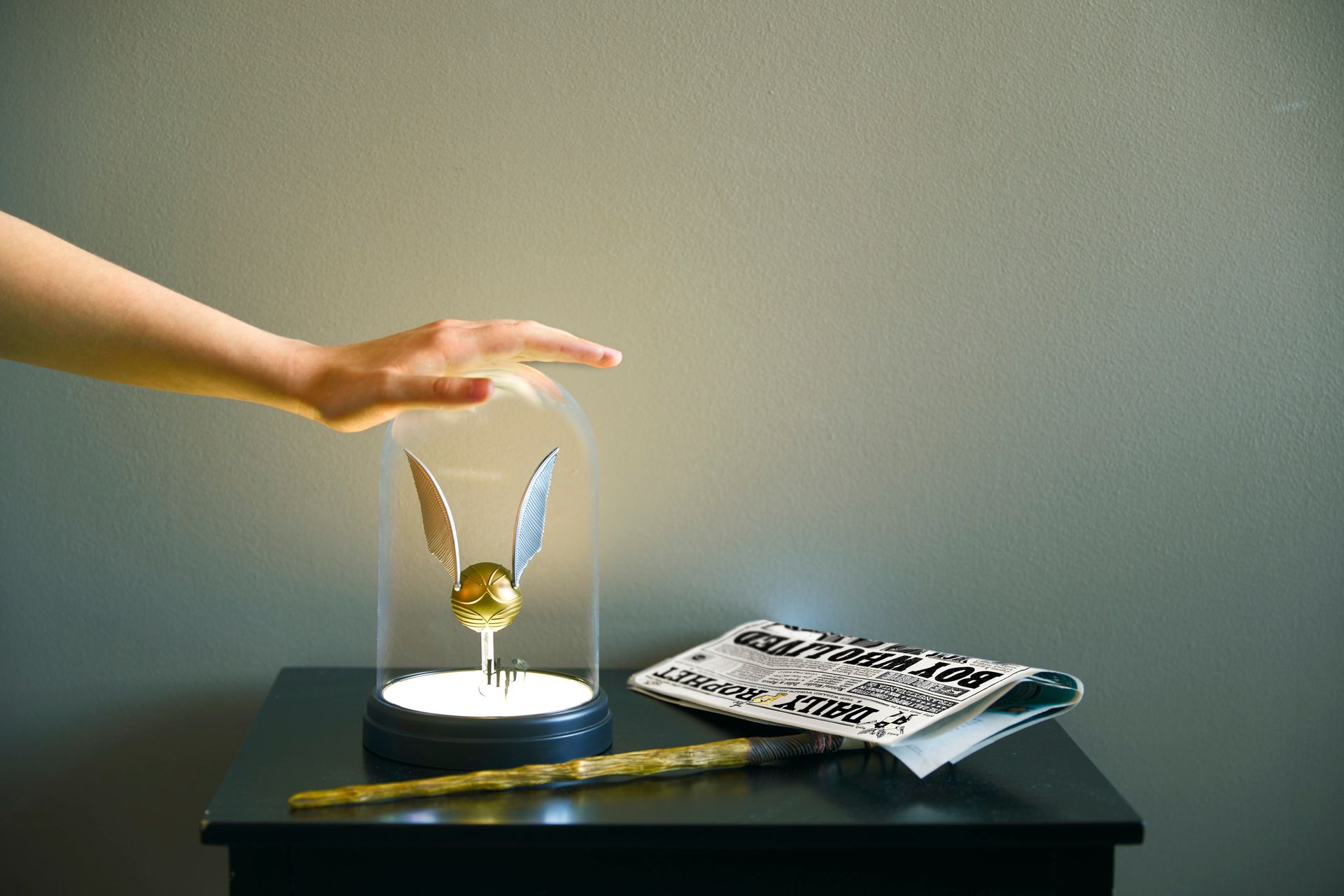 A person is touching a golden snitch in a glass dome on a table.