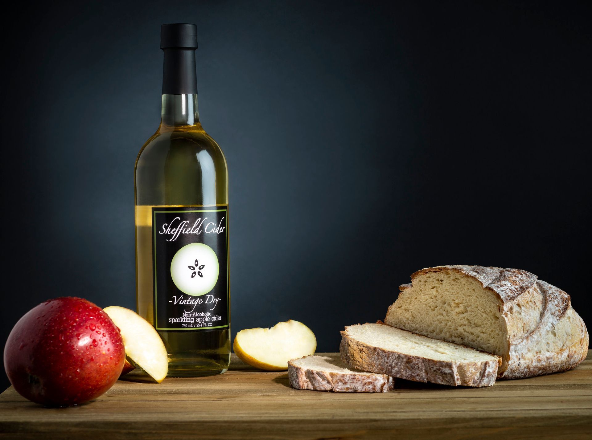 A bottle of apple cider sits on a wooden table next to apples and bread.