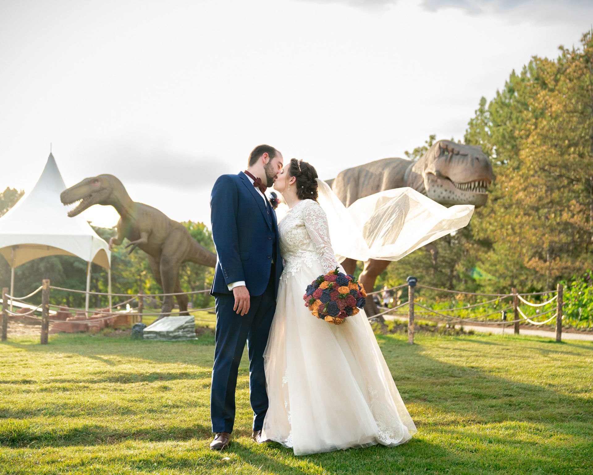 A bride and groom are kissing in front of a field of dinosaurs.