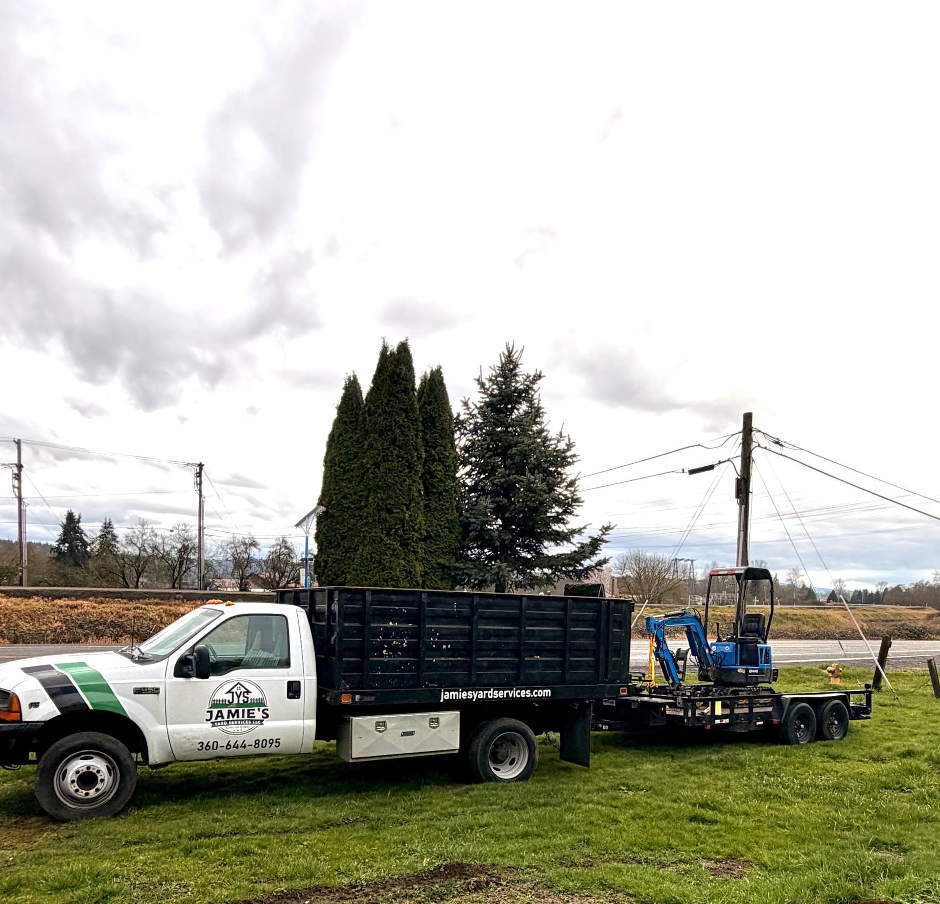 An excavator on a cleared lot with a house and trees in the background under an overcast sky.