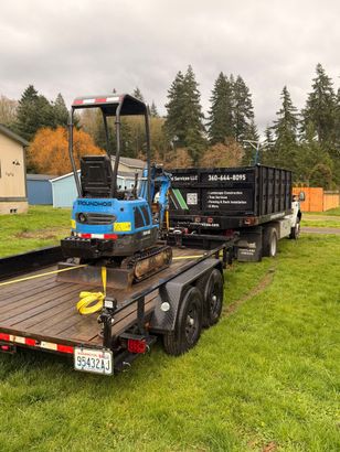 Mini excavator on a trailer next to a dump truck; green grass, trees and a cloudy sky are in the background.