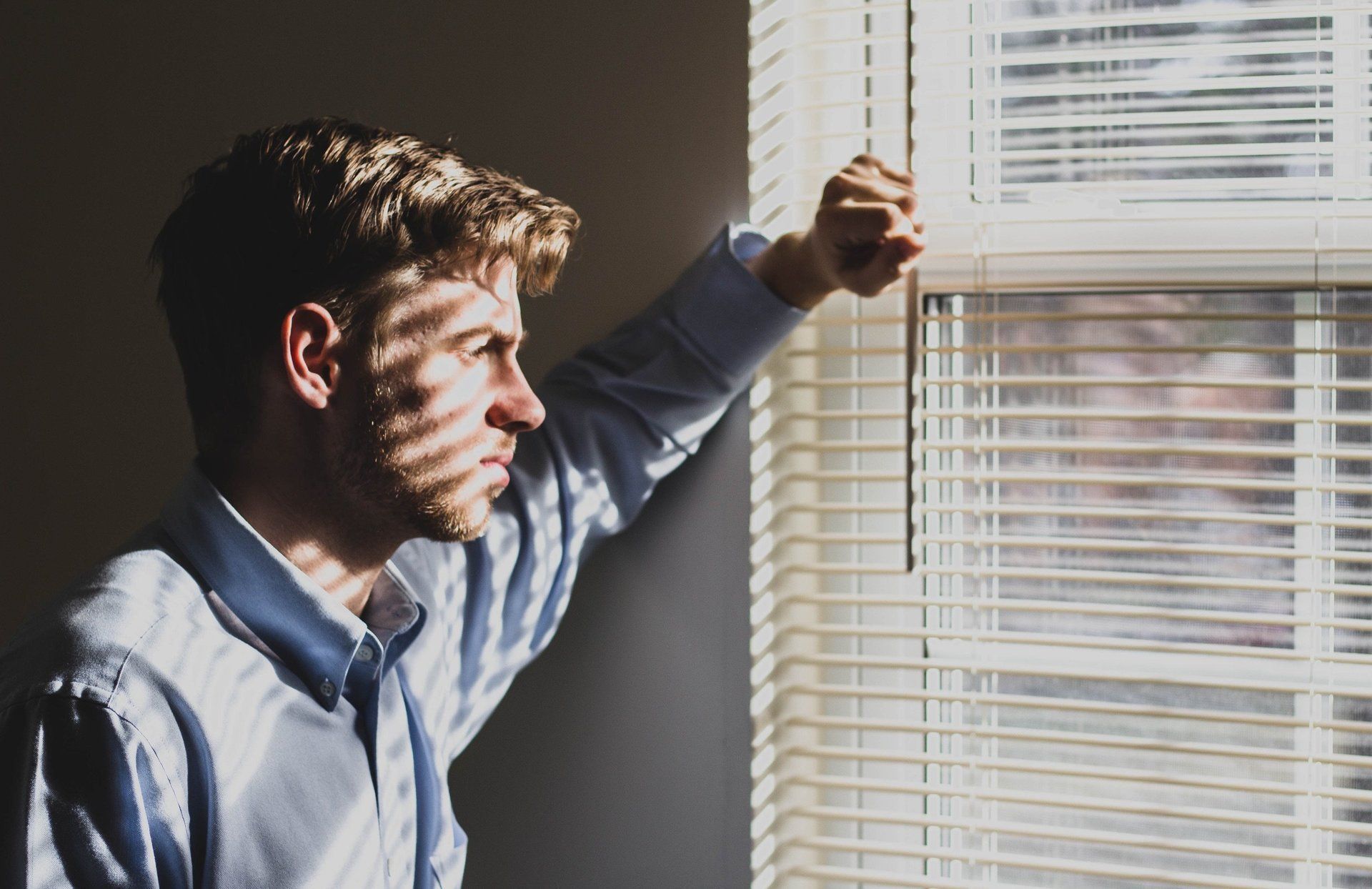 A man is looking out of a window with blinds.