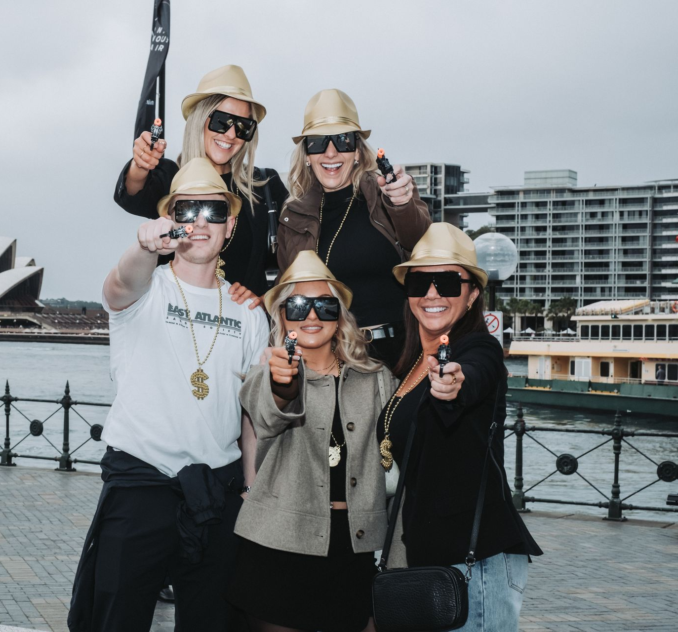 Five friends in straw hats and sunglasses pose together by a waterfront railing, smiling and pointing at the camera.