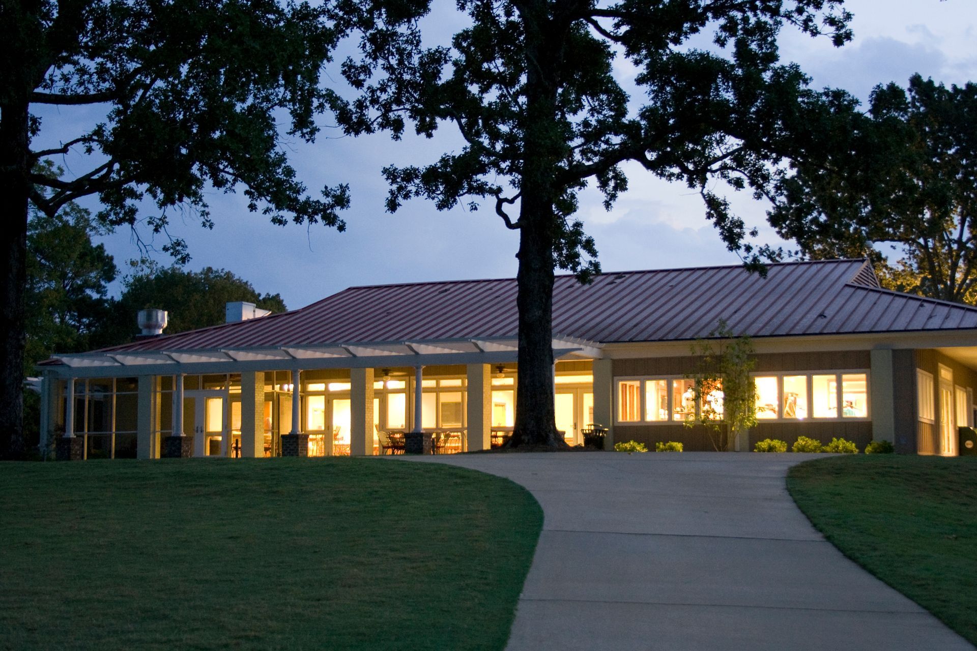 Pathway leading to a lit-up building with a red roof at dusk, framed by trees and green lawn.