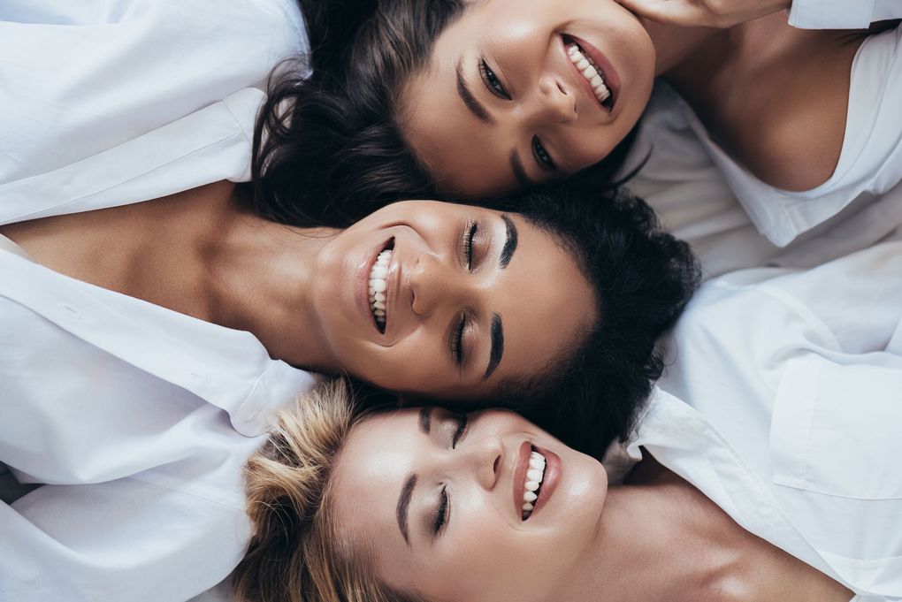 Three women are laying on a bed with their heads together and smiling.