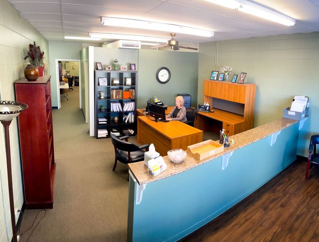 A woman sits at a desk in an office with a clock on the wall