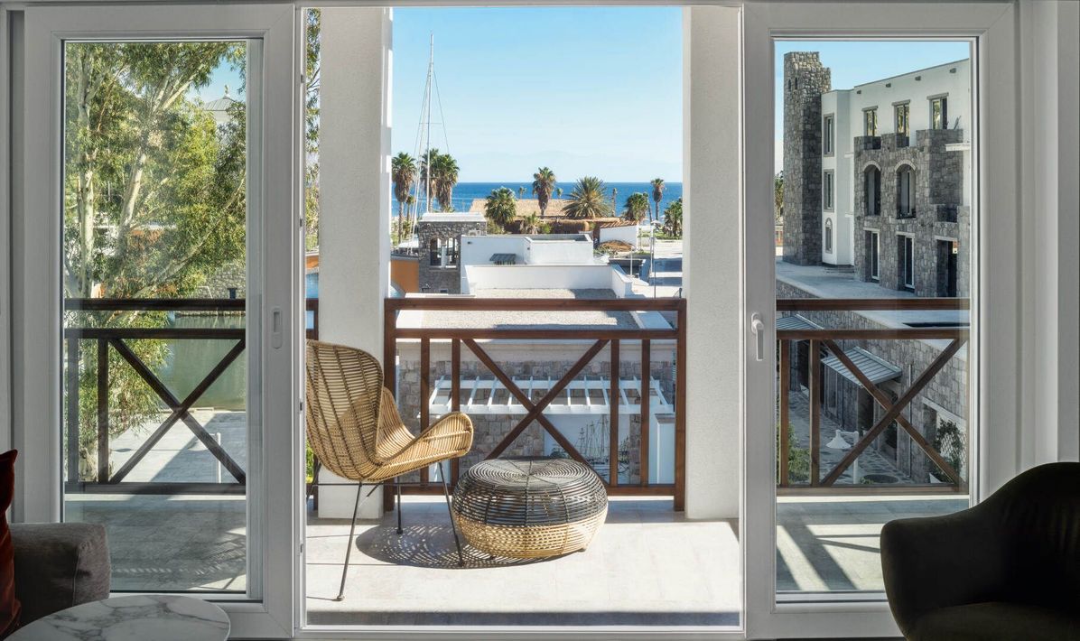 Vista dal balcone attraverso le porte aperte su un lungomare soleggiato con sedia, pouf e edifici sullo sfondo.