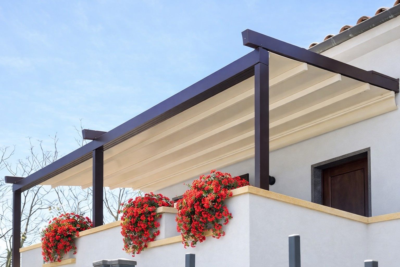 Terrazza di una casa moderna con tenda da sole color crema, travi in ​​legno scuro e piante rampicanti con fiori rossi su un muro bianco.