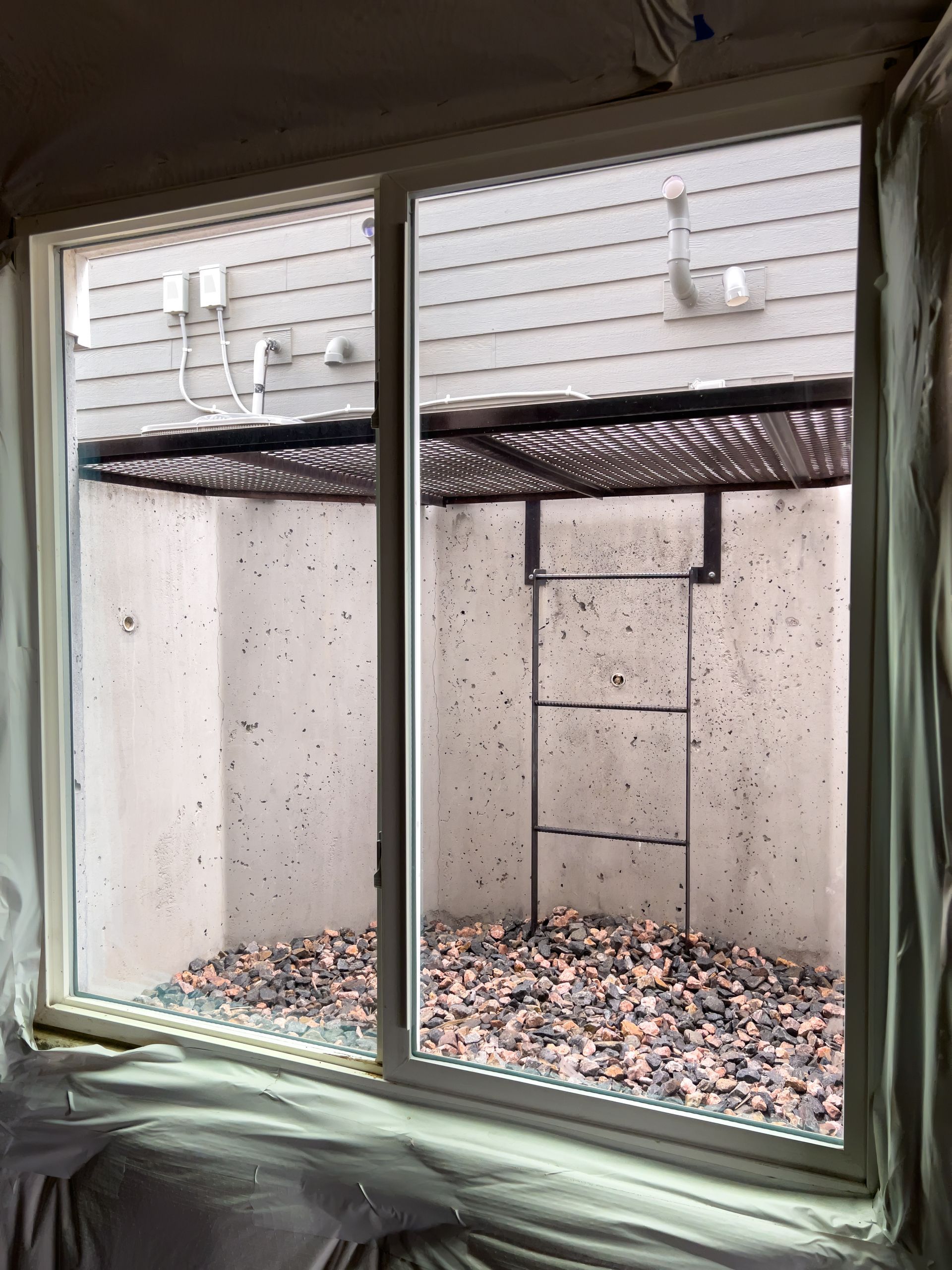 White-framed sliding window with view of a sunken window well filled with gravel and a ladder.