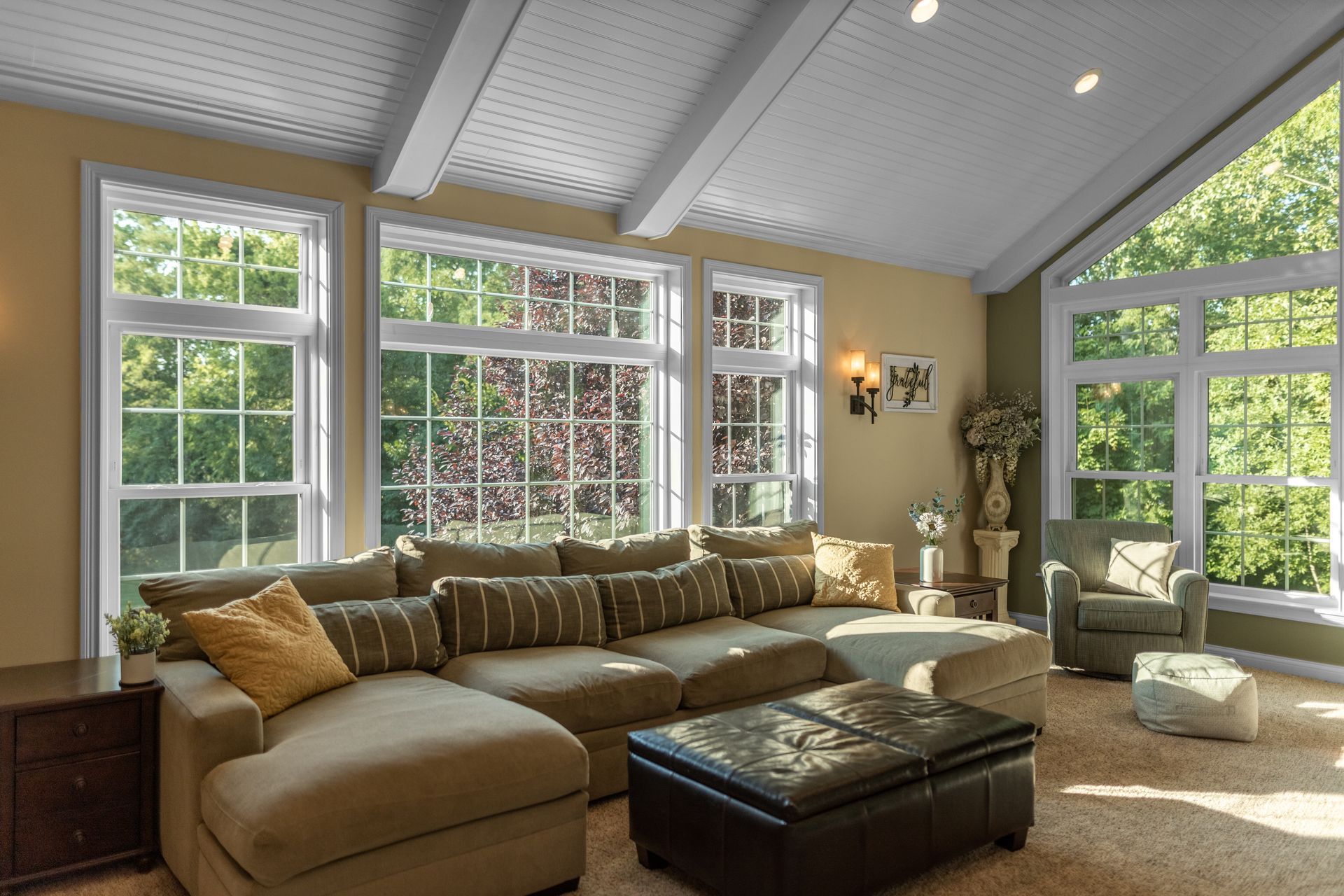 Living room with large windows, sectional sofa, and ottoman. Beige walls and white ceiling.
