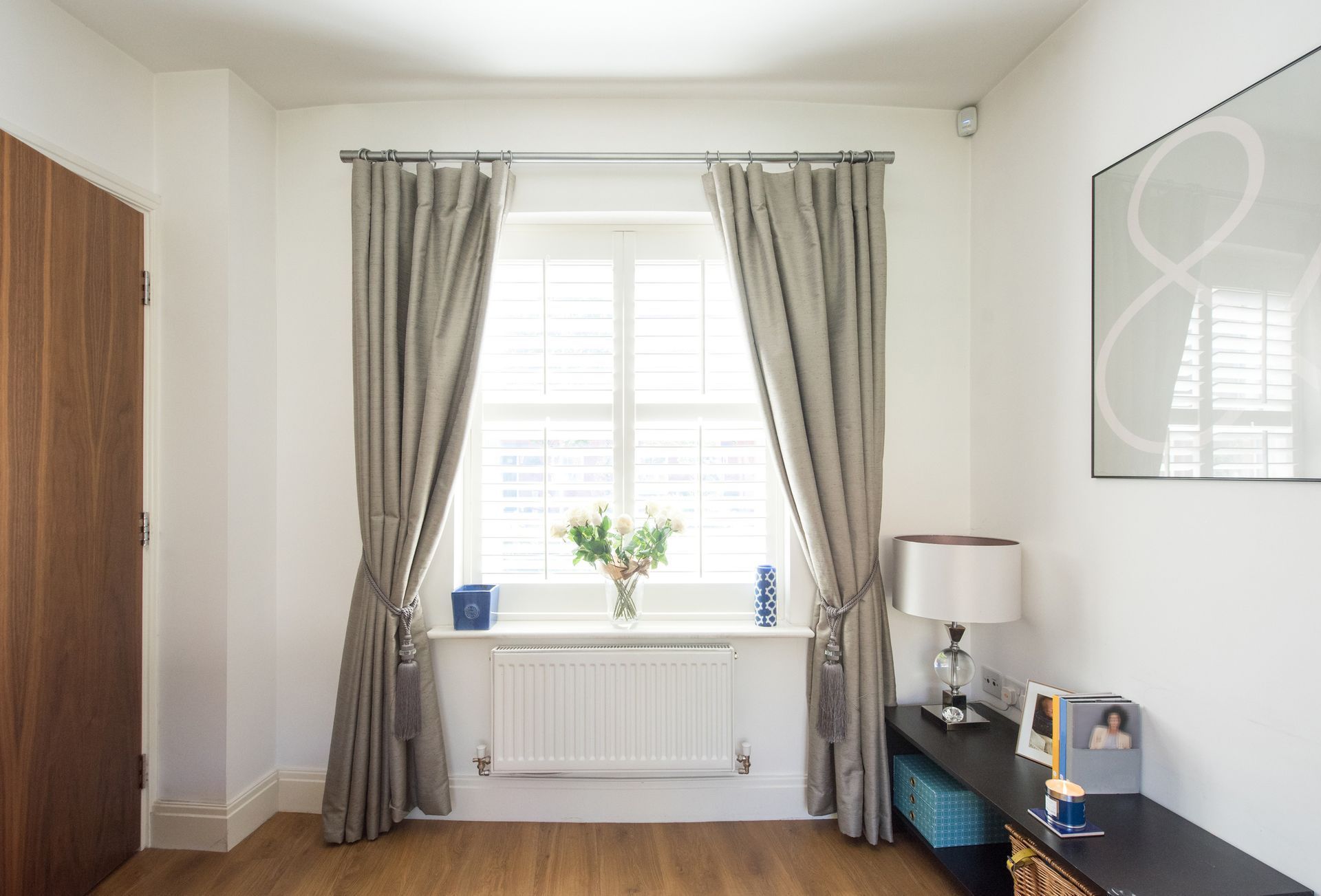Room interior with window, curtains, and radiator. Gray curtains tied back, white walls, and wood floor.