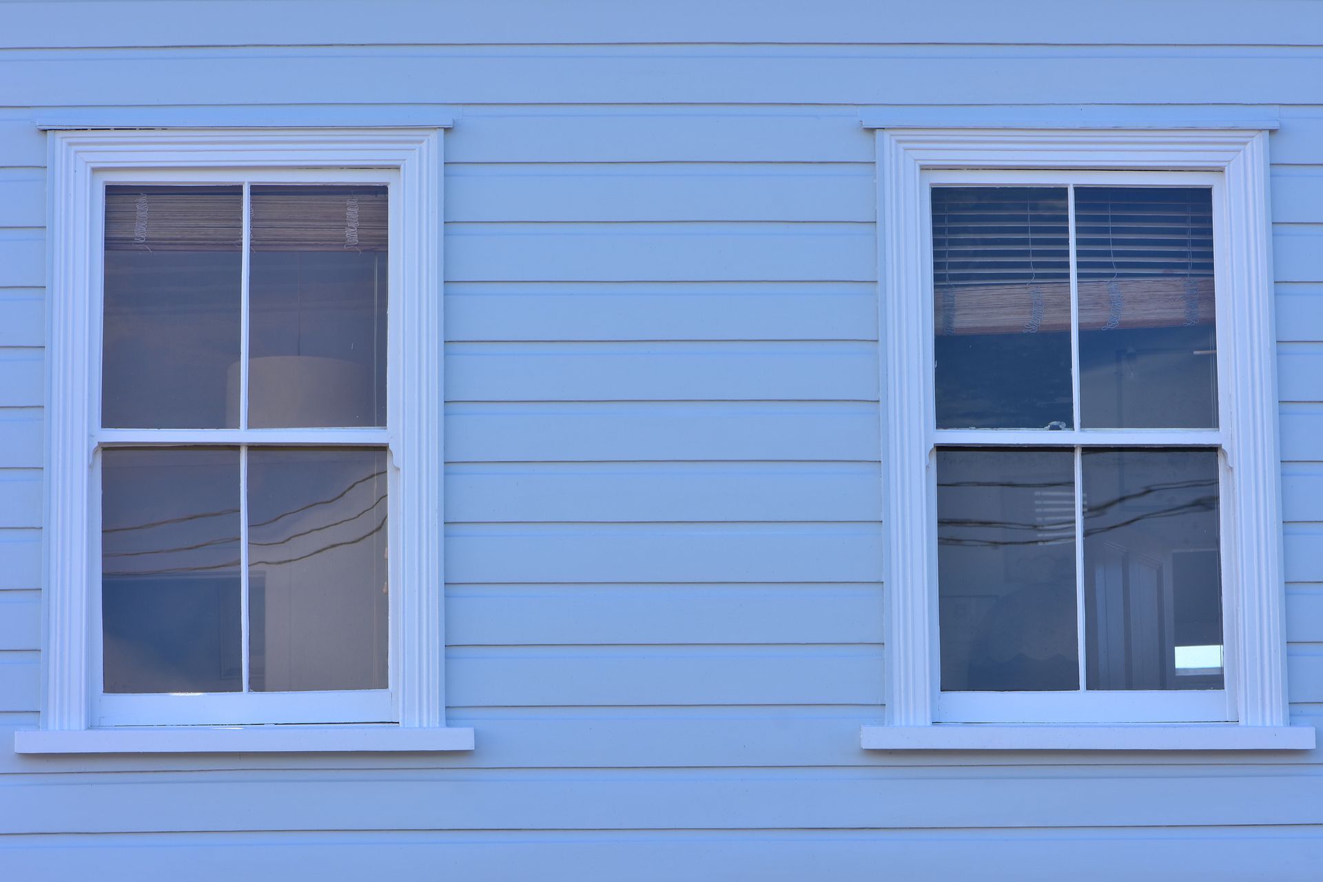 Two white-framed windows on a light blue clapboard wall. One window reflects a clear view, the other has blinds. Two white-framed windows on a light blue clapboard wall. One window reflects a clear view, the other has blinds.