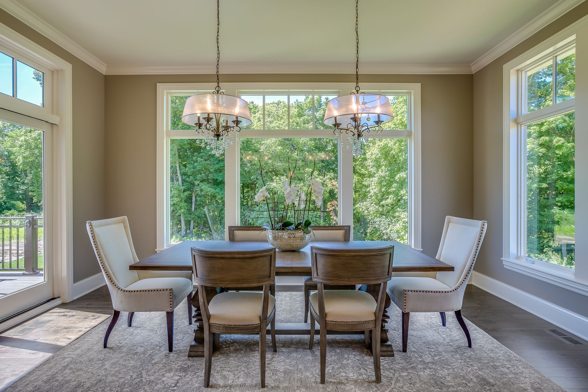 Dining room with rectangular table, six chairs, two chandeliers, and large windows with greenery views. Dining room with rectangular table, six chairs, two chandeliers, and large windows with greenery views.