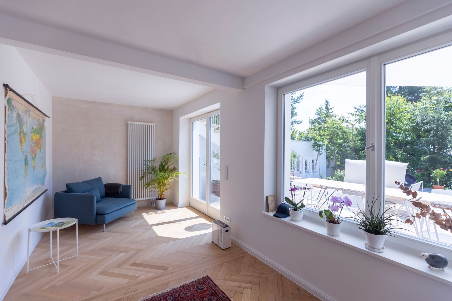 Bright living room with blue armchair, large windows overlooking greenery, and a world map. Bright living room with blue armchair, large windows overlooking greenery, and a world map.