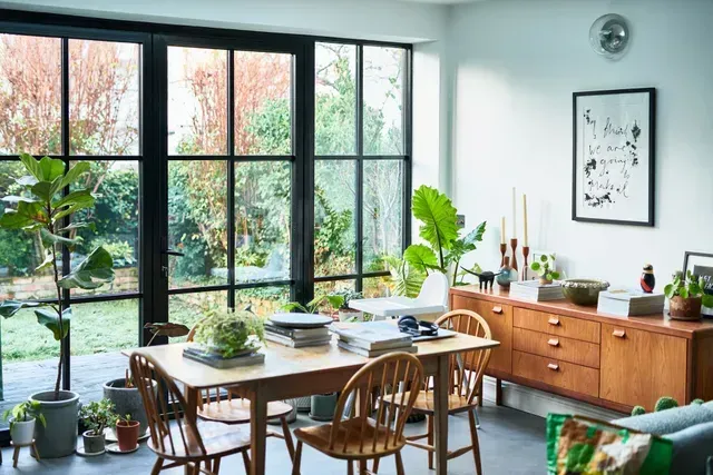 Dining room with large window overlooking garden, table, chairs, plants, and wooden sideboard. Dining room with large window overlooking garden, table, chairs, plants, and wooden sideboard.
