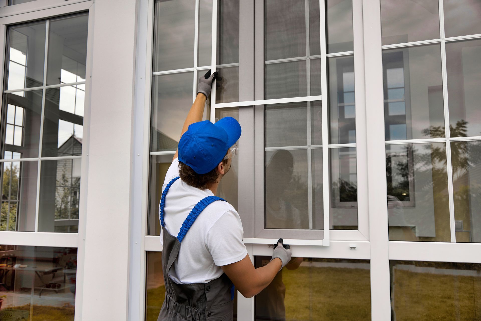 Person in overalls and blue cap installing a window screen on a white framed window. Person in overalls and blue cap installing a window screen on a white framed window.