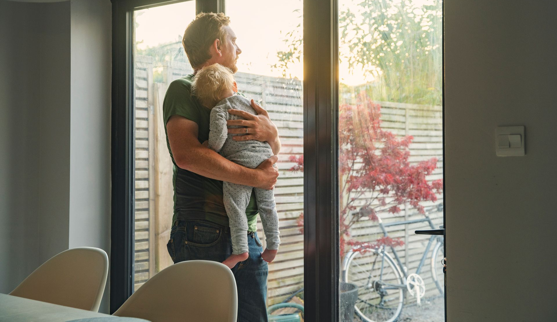 Man holding a child, looking out a window at a garden with a bicycle. Man holding a child, looking out a window at a garden with a bicycle.