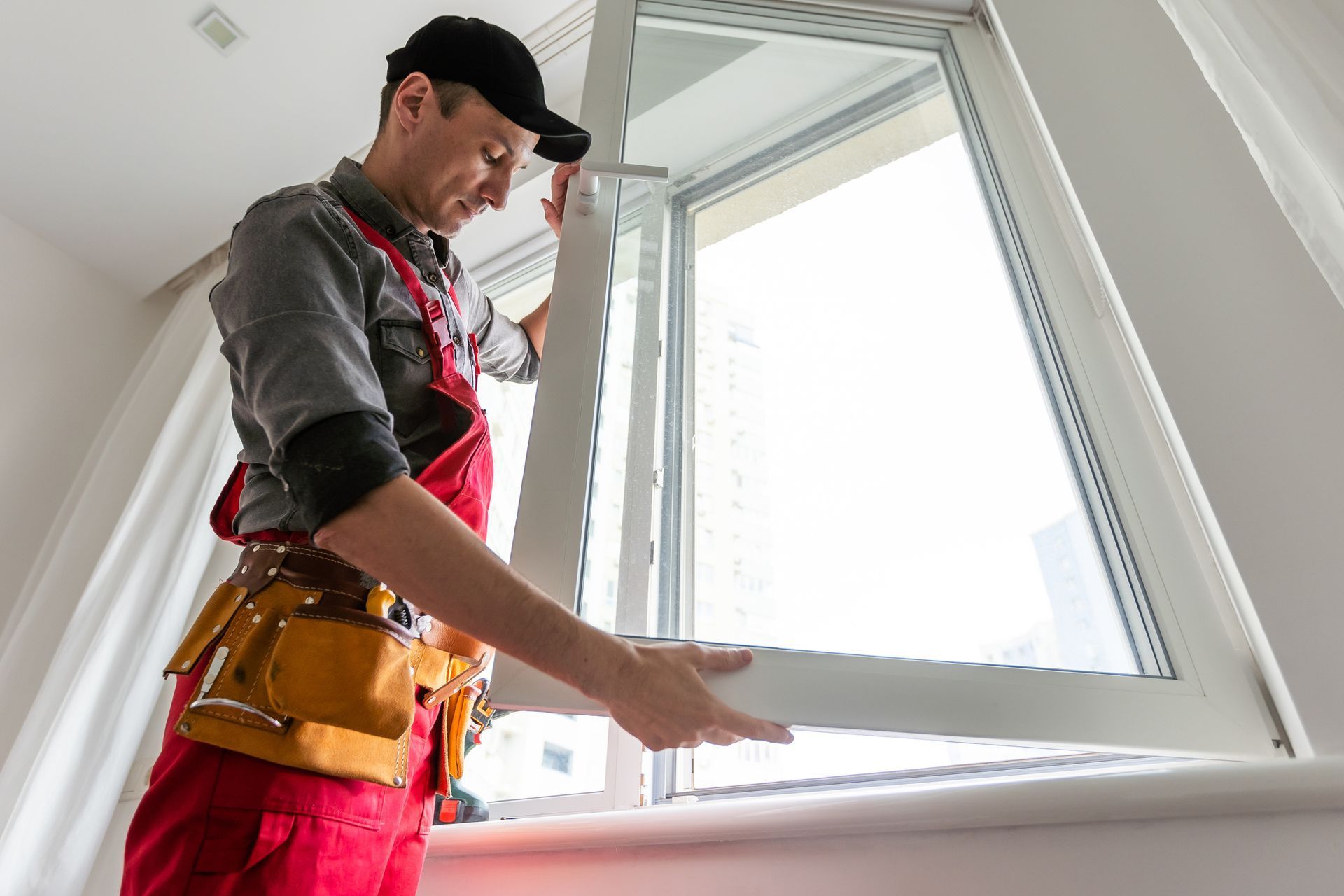 Person in red overalls and black cap installing a window, indoors. Person in red overalls and black cap installing a window, indoors.