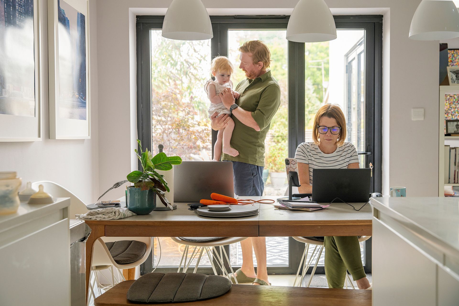 Family working at home: parent holding child, another working on a laptop, sunny room with open doors.