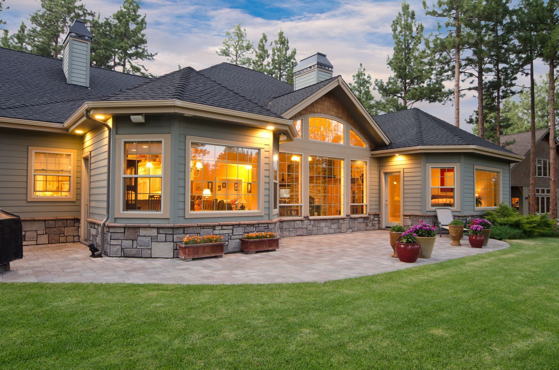 House exterior with patio, illuminated windows, and lawn. Green siding, stone accents, and surrounding trees.