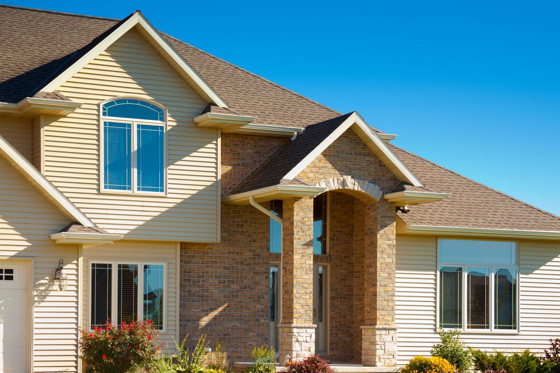 Beige and brick home with white trim and a brown roof against a blue sky.