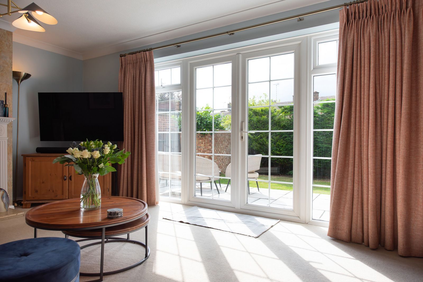 Living room with pink curtains, a round coffee table, and French doors leading to a garden.