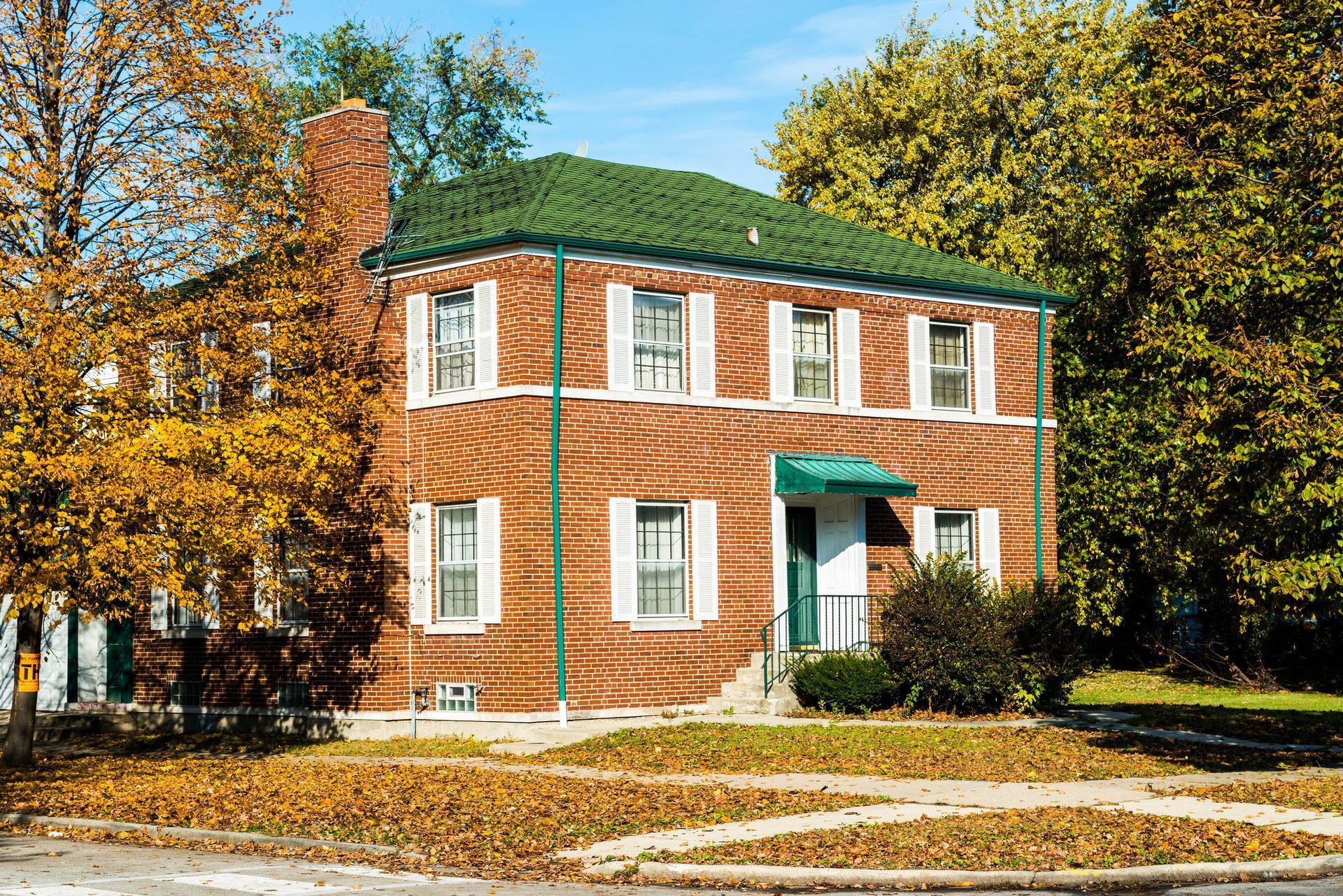 Two-story brick house with green roof, white shutters, and fall foliage. Two-story brick house with green roof, white shutters, and fall foliage.