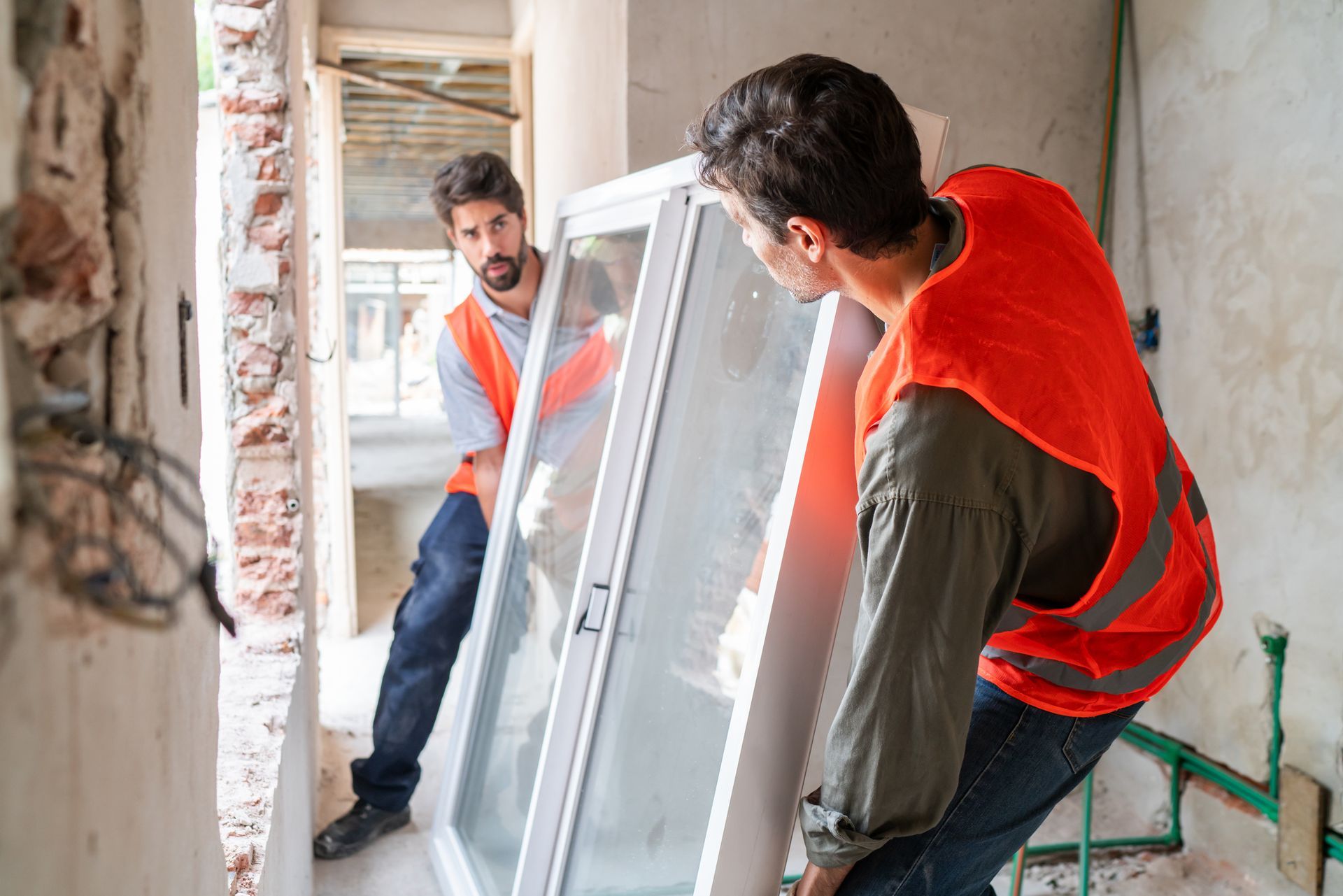 Two workers carry a large window through a construction site hallway, both wearing orange vests.