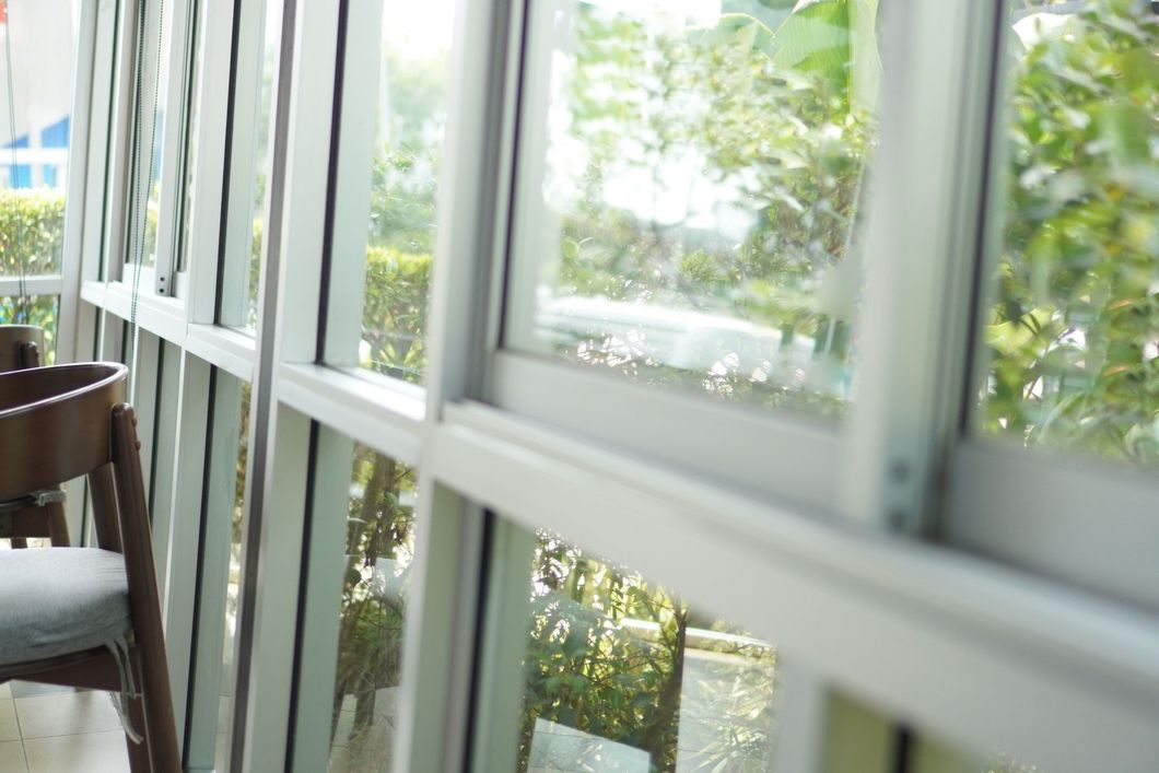 White-framed windows overlooking greenery. Wooden chair visible on the left side. Sunny daylight. White-framed windows overlooking greenery. Wooden chair visible on the left side. Sunny daylight.