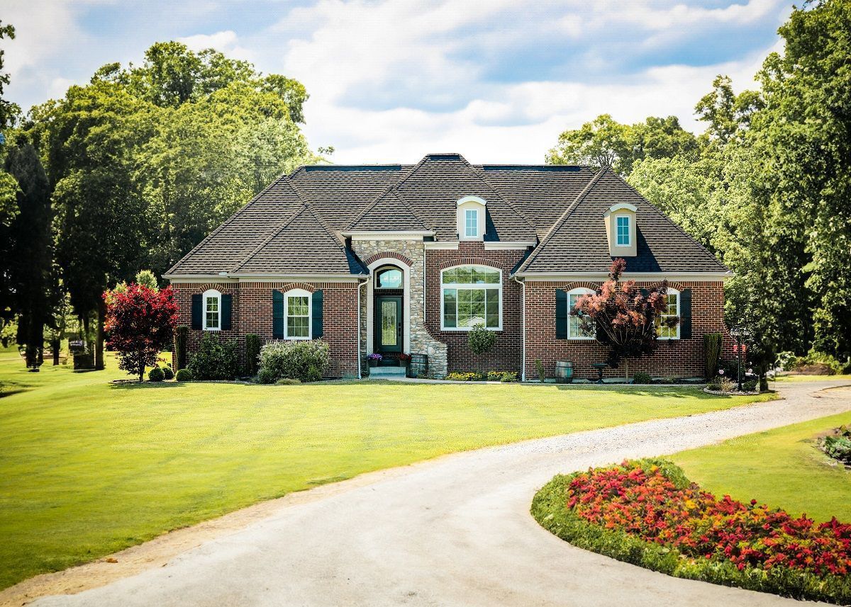 Brick house with green shutters, a tiled roof, and a winding driveway on a well-manicured lawn surrounded by trees. Brick house with green shutters, a tiled roof, and a winding driveway on a well-manicured lawn surrounded by trees.