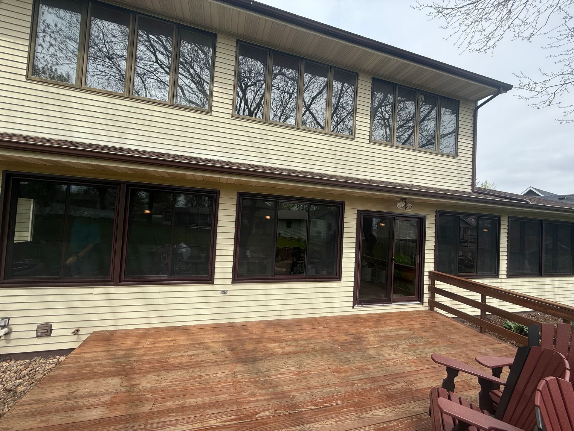 Beige house with brown-framed windows, overlooking a weathered wooden deck with Adirondack chairs.