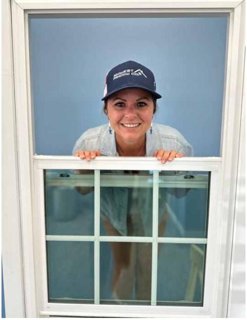 Woman peeking through a window, wearing a hat, smiling. Blue wall behind her.