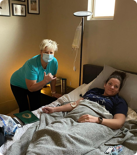 A woman wearing a mask is standing next to a woman laying in a bed.
