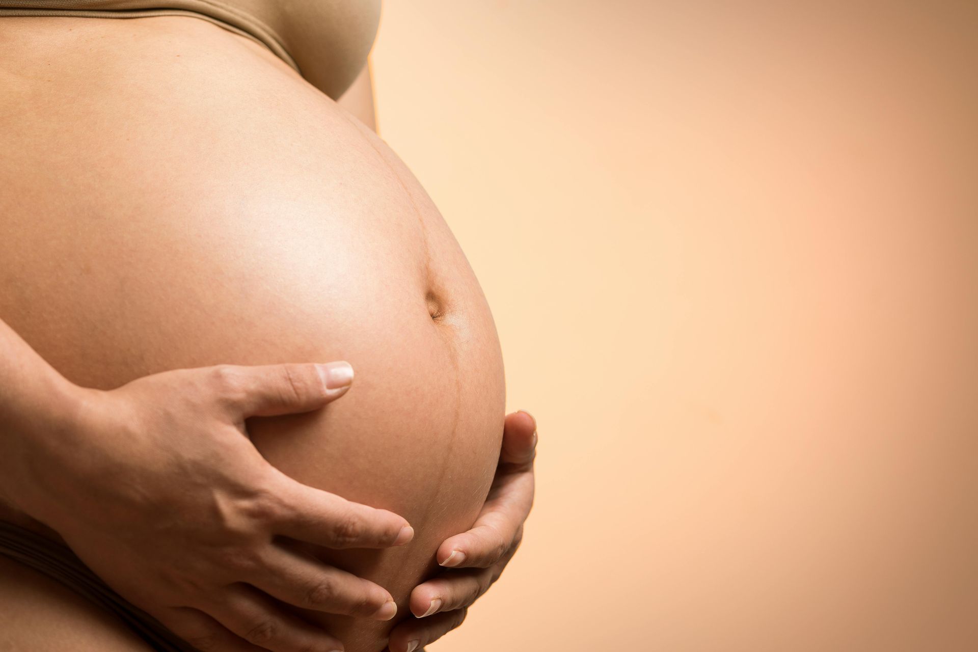 Pregnant woman's hands cradling her prominent belly, set against a warm, neutral background.