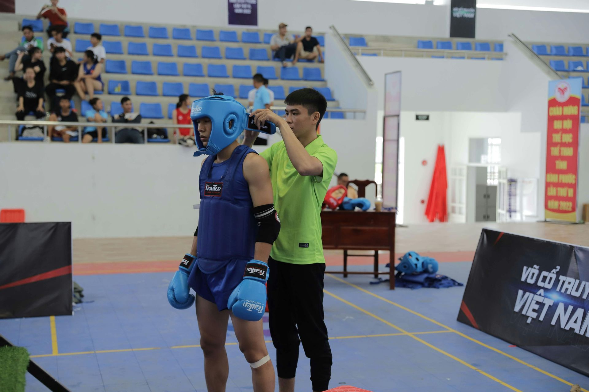 A fighter in blue gear gets his headgear adjusted by a man in a green shirt in an arena.