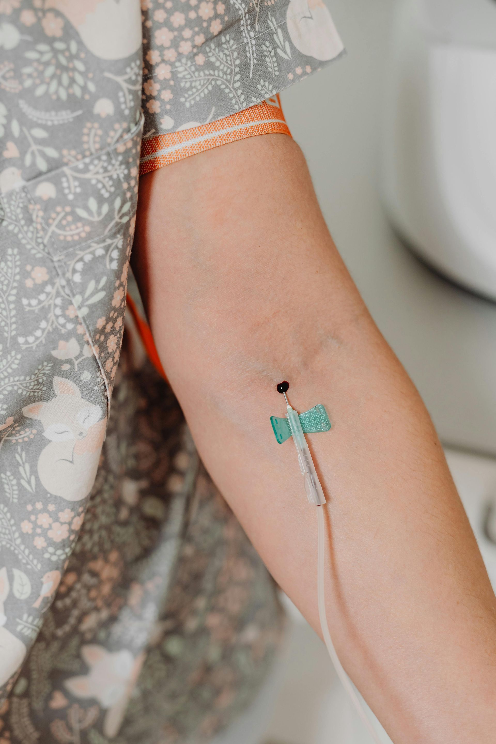 Arm with an IV needle inserted, medical setting. The person wears a floral patterned shirt.