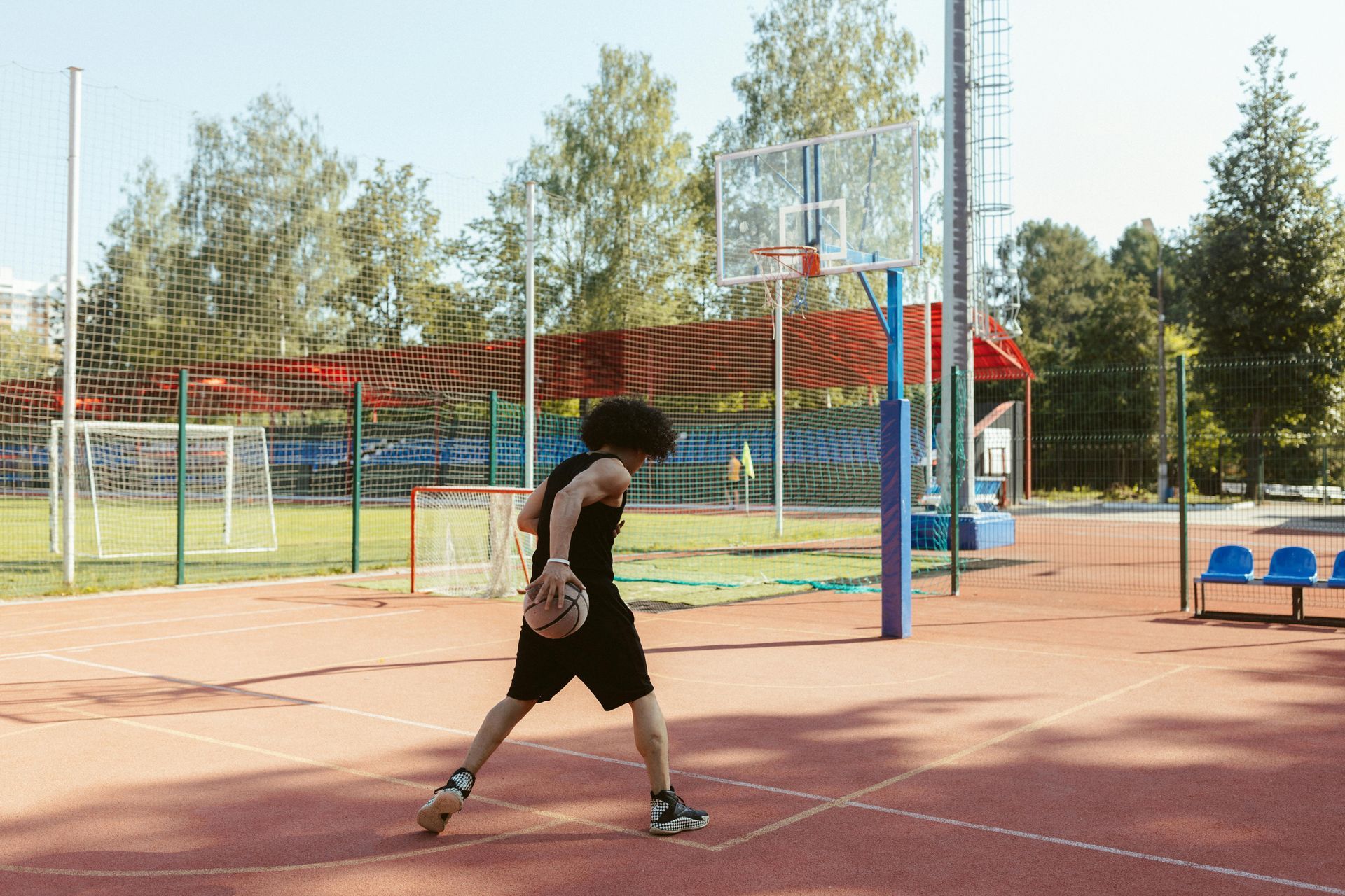 A person in black shorts and tank top dribbles a basketball on an outdoor court with a hoop.
