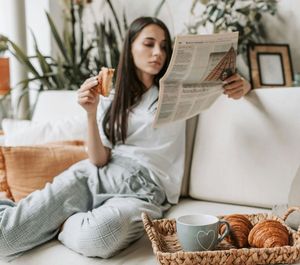 A woman is sitting on a couch reading a newspaper and eating croissants.