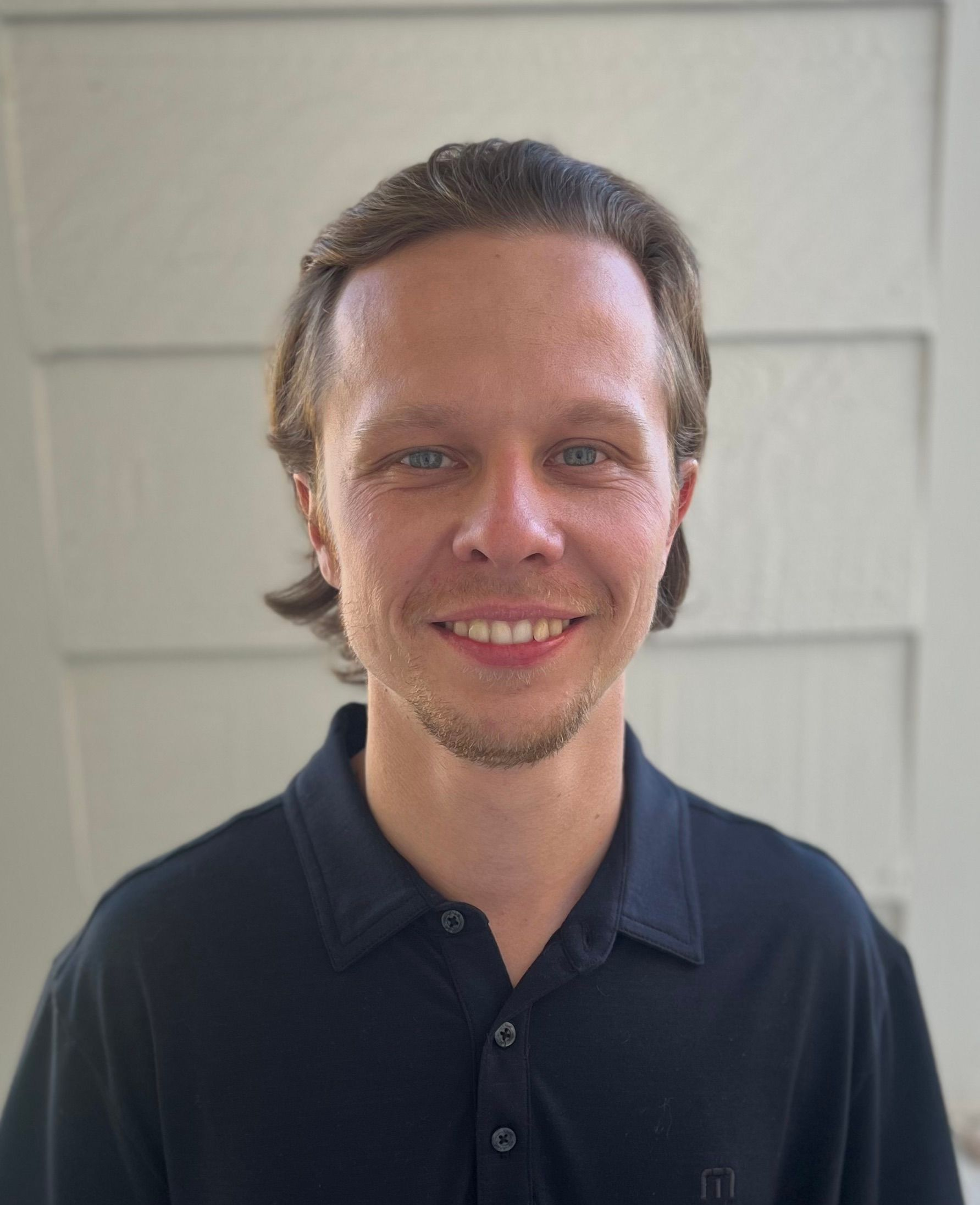 Man smiling outdoors, wearing a hooded sweatshirt. Sunlight, house, and bare branches in the background.