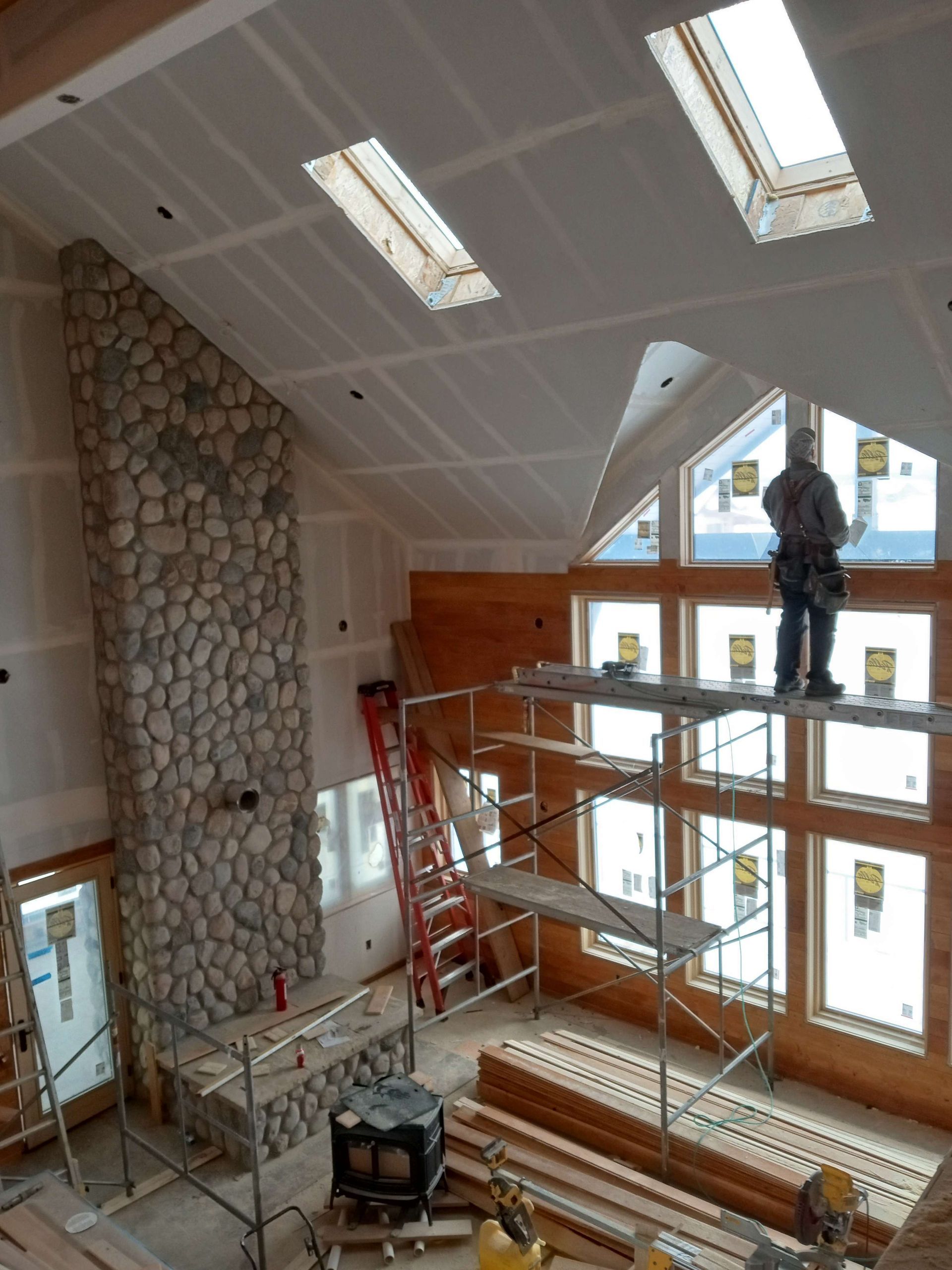 Construction site with stone fireplace, skylights, a worker on scaffolding, and lumber.