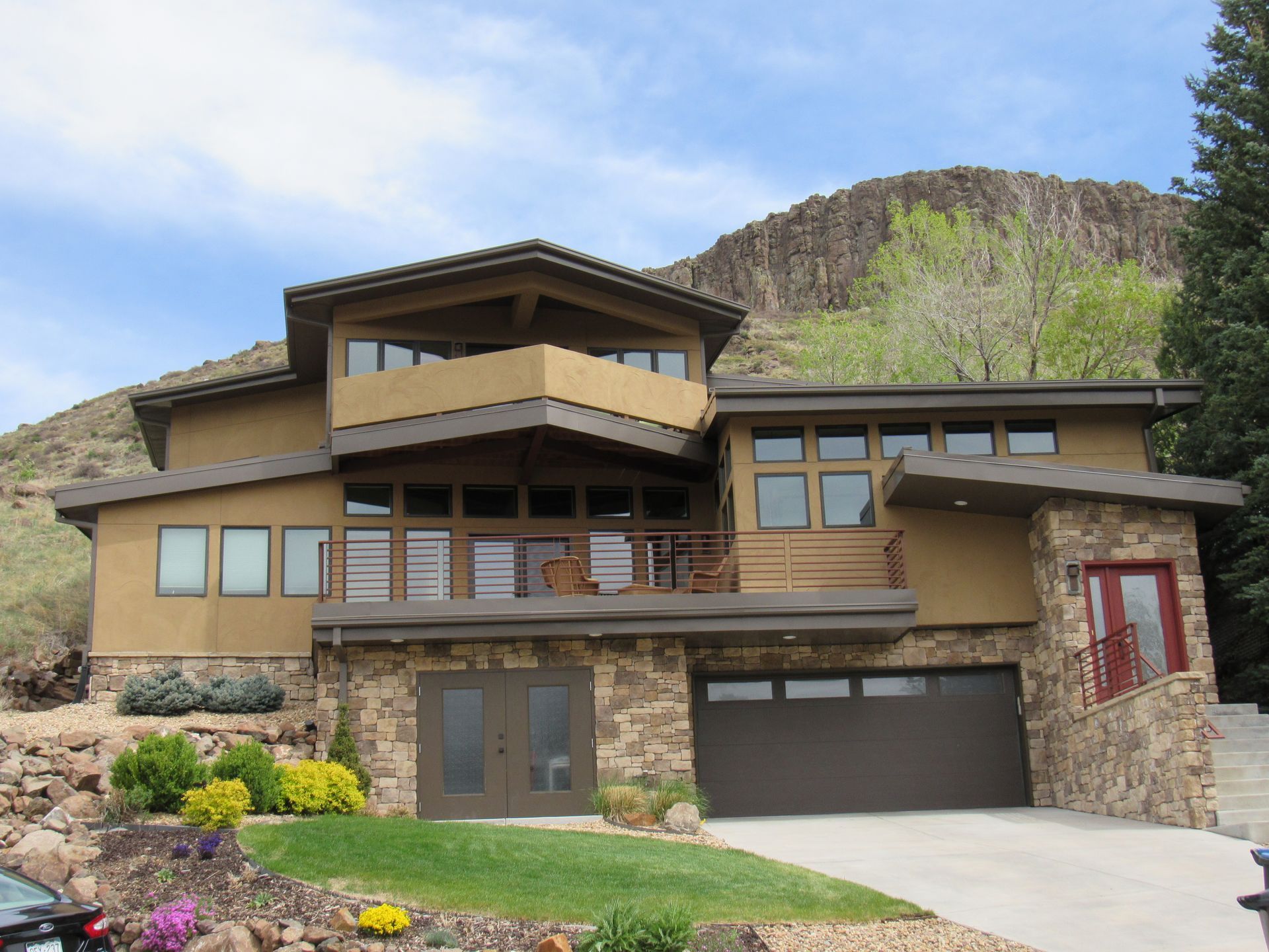Two-story house with tan stucco, brown trim, stone accents, and mountain backdrop.