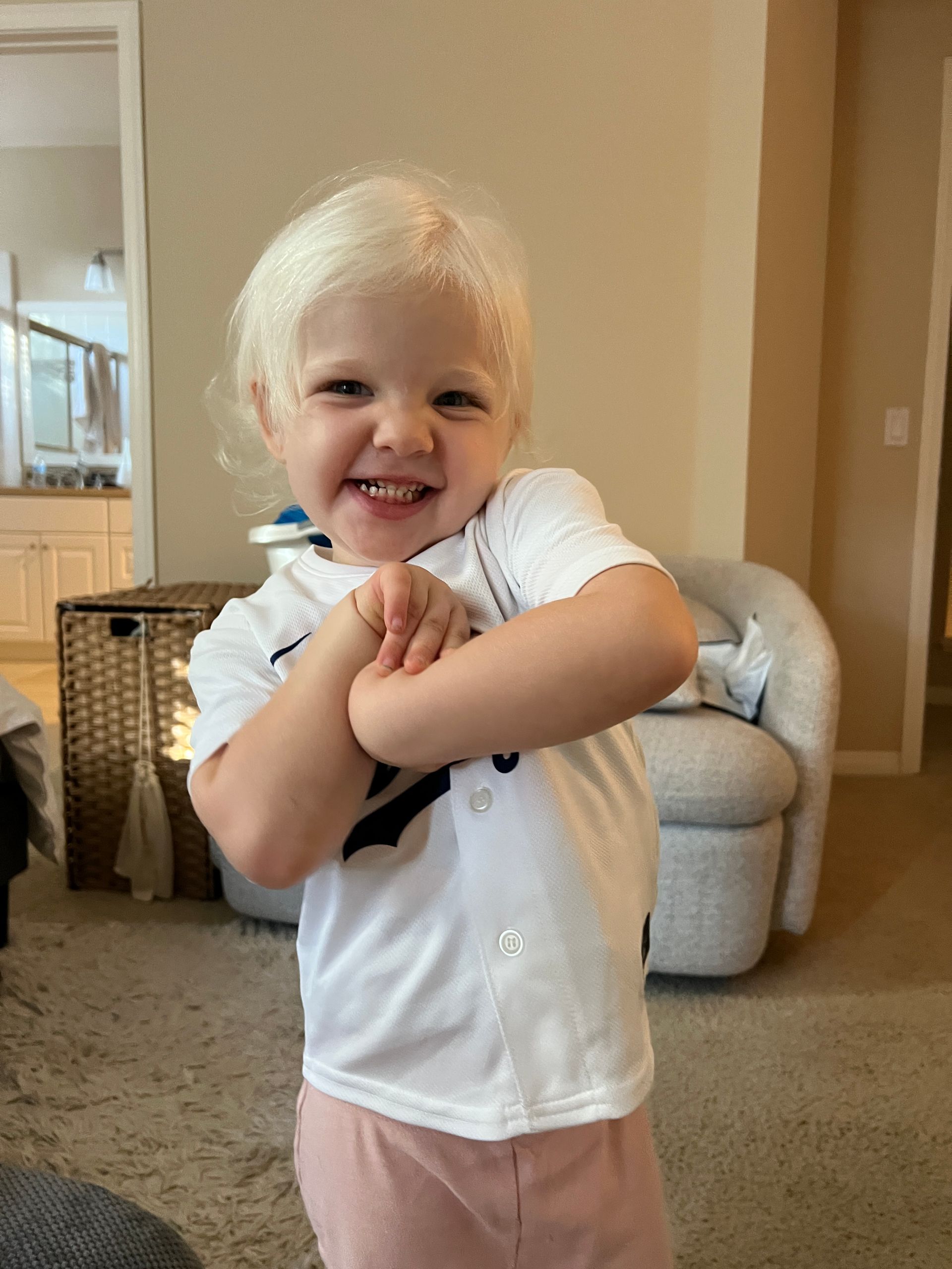 Blond-haired child in white jersey and pink pants smiles with arms crossed, in a living room.