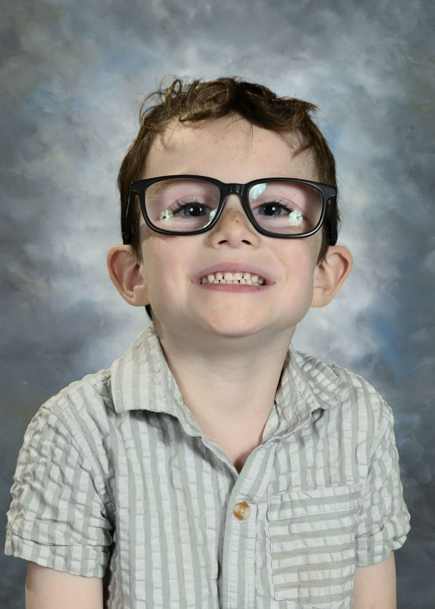 Young child with glasses, wearing a white shirt with patches, indoors.