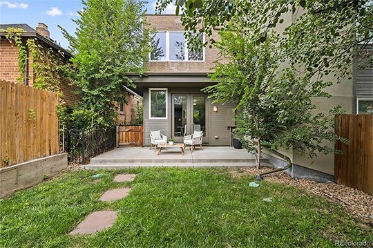 Backyard with patio, chairs, small table, lawn, and two-story house in background. Trees and fences flank the yard.