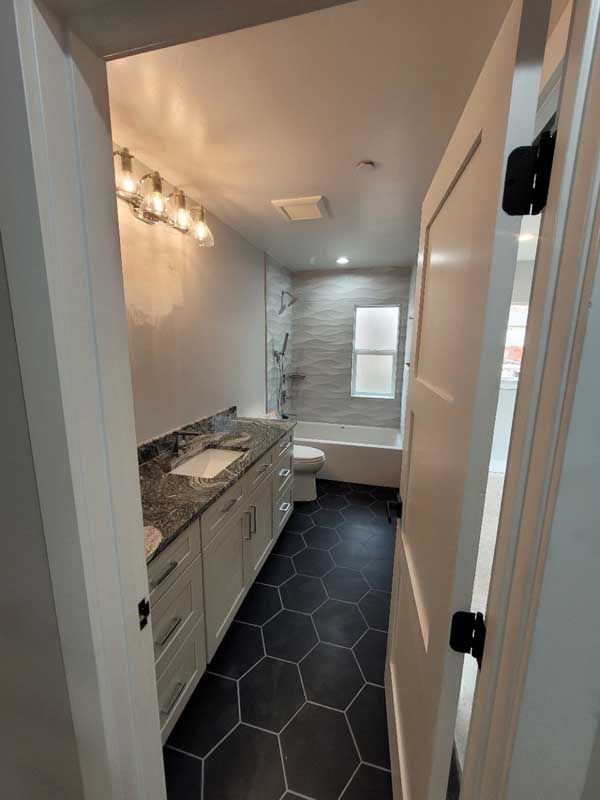 Bathroom with white vanity, black hexagon floor tiles, gray tile shower, and a white door.