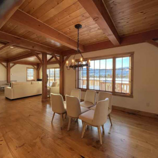 Spacious dining room with wooden beams, light wood floor, and six beige chairs. Large window overlooks snowy mountains.