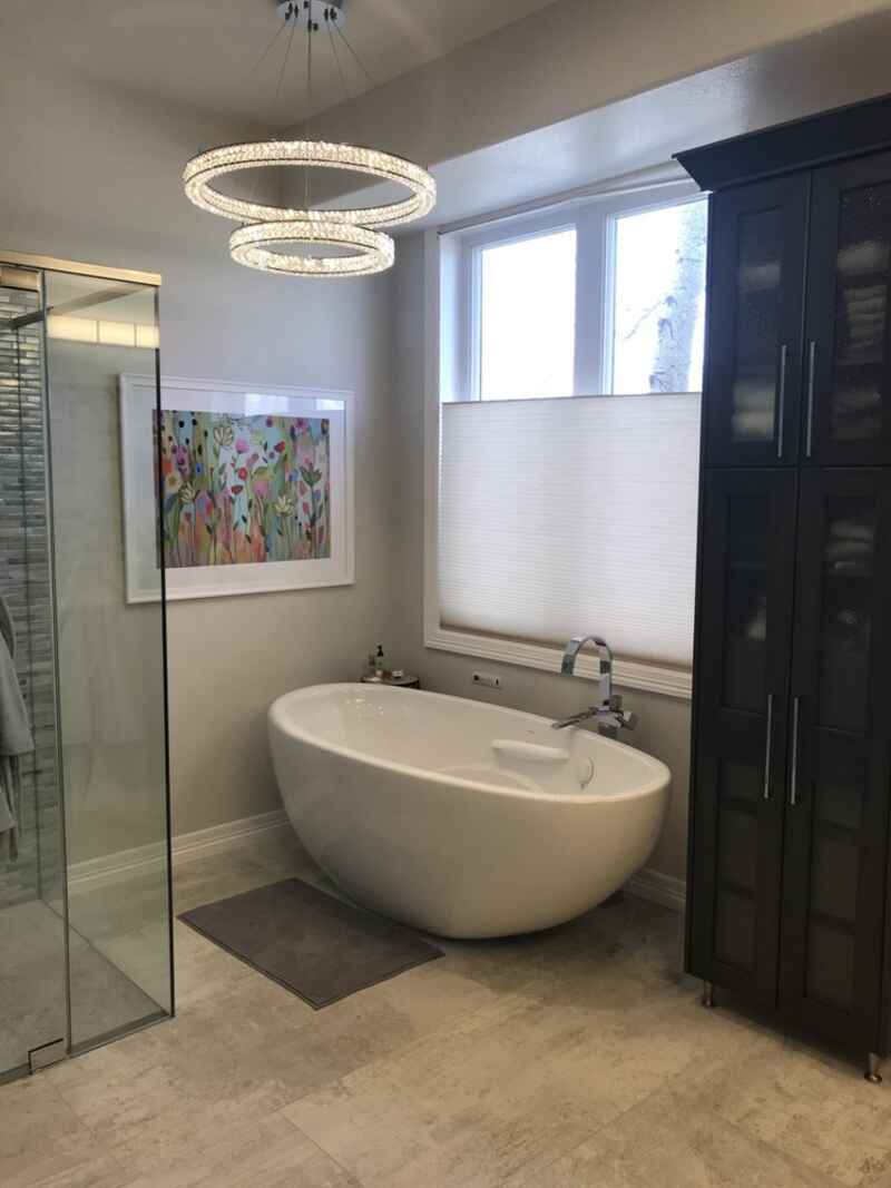 Bathroom with a white oval tub, art, dark storage cabinet, and a window with a shade.