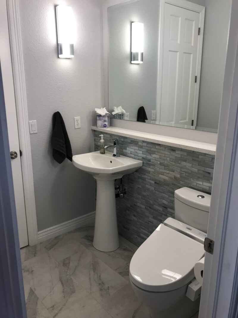 Small bathroom with pedestal sink, toilet, gray tile wall, and bright sconces.