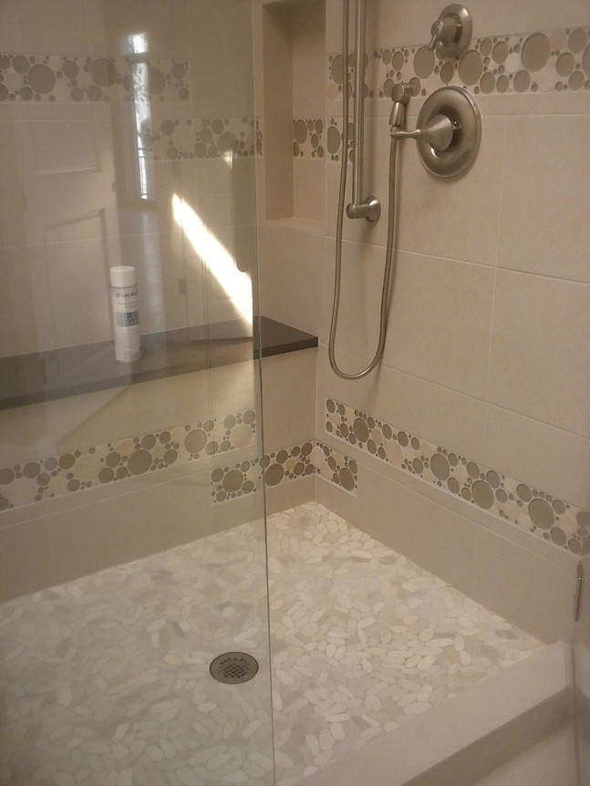 Shower with white pebble floor, glass door, and built-in shelf. Beige tile with floral pattern border.